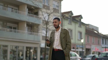 Düşünceli bir Orta Doğulu adam derin düşünce ifadesiyle elinde telefonla caddede dikiliyor. Düşünceli bir Orta Doğu kararı ve teknolojiyi kullanarak