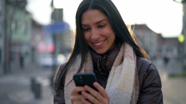 Şehir caddesinde telefona bakan mutlu bir Orta Doğulu genç kadın. 20 'li yaşlarda bir kadın, şehir merkezinde elinde teknoloji cihazıyla dikiliyor.