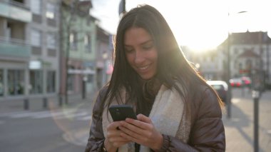 Gün batımında güneş ışığıyla parlayan bir cep telefonu kullanan mutlu genç bir kadın. 20 'li yaşlarda elinde teknoloji cihazı olan bir kadın ekrana bakıyor.