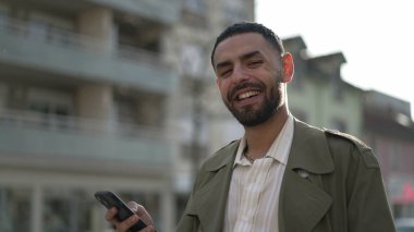 Mutlu genç adam gülüyor ve yakından gülümsüyor. Şehir caddesinde duruyor. Orta Doğu kökenli bir Arap erkeği gülüyor ve gülümsüyor.