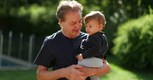 Grand-father holding baby infant in arms outside in backyard. Grand parent bonding with grand-child