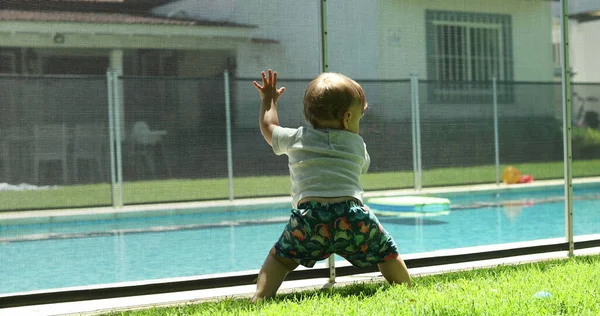 Infant baby leaning on pool fence with ball. Toddler child playing outside during summer day
