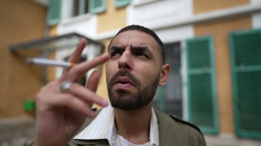 Düşünceli bir Orta Doğulu genç adam dışarıda sigara içiyor düşünceli bir ifadeyle. Düşüncelere dalarken yakın çekim görüntüsü alınıyor