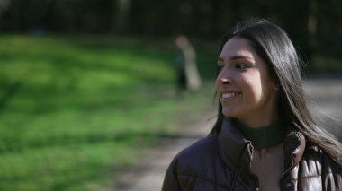 Kendinden emin genç bir kadın güneşli bir günde parkta yürüyor. Mutlu kadın yüzü kapalı yürüyüp güneşleniyor ve temiz hava alıyor.