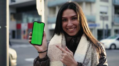 Neşeli genç bir kadın telefonda şehir caddesinde krom renkli yeşil ekran sunuyor. Kameraya çevrimiçi video gösterilirken akıllı telefon ekranını gösteren bir kadın.