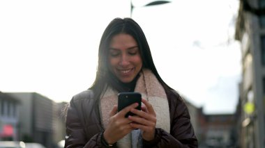 20 'li yaşlarda, elinde cep telefonu olan mutlu bir yetişkin kız. Şehir merkezinde telefondaki mesajları okuyan genç bir kadının hareket halindeki görüntüsü.