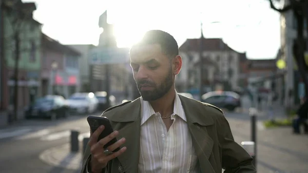 自信を持っている中東の男性の肖像午後の日差しのフレアの間に街の通りの外に立っている間 電話画面を見て — ストック写真