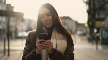 Gün batımında şehir sokaklarında yürürken elinde cep telefonu tutan mutlu bir Orta Doğulu genç kadın.