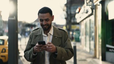 Mutlu bir Orta Doğulu genç adam güneşli bir günde elinde akıllı telefon cihazıyla şehirde yürüyor.