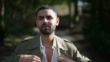 Güneşli bir günde parkın dışında yürüyen ve ceketini ayarlayan kendinden emin genç bir Arap. Modaya uygun mutlu insan.