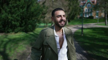 Neşeli genç bir Arap adam güneşli bir parkta güneşli bir şekilde yürüyor, güneşin sıcaklığında güneşleniyor ve dışarıdaki güzelliğin tadını çıkarıyor.