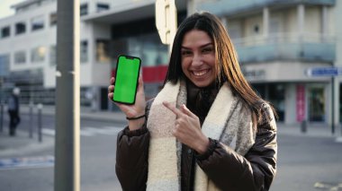 Neşeli genç bir kadın telefonda şehir caddesinde krom renkli yeşil ekran sunuyor. Kameraya çevrimiçi video gösterilirken akıllı telefon ekranını gösteren bir kadın.