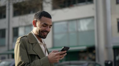 Bir mutlu Arap genç adam, elinde cep telefonu cihazıyla caddede dururken gülümsüyor. Gülümseyen ve teknolojiyi kullanan Orta Doğulu bir erkeğin yakından çekilmiş yüzü.