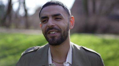 Güneşli bir günde parkta gülümseyen, kameraya doğru yürüyen mutlu bir Arap adam.