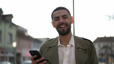 Bir mutlu Arap genç adam, elinde cep telefonu cihazıyla caddede dururken gülümsüyor. Gülümseyen ve teknolojiyi kullanan Orta Doğulu bir erkeğin yakından çekilmiş yüzü.