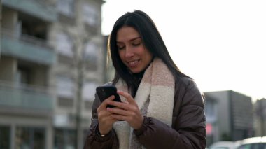 20 'li yaşlarda, elinde cep telefonu olan mutlu bir yetişkin kız. Şehir merkezinde telefondaki mesajları okuyan genç bir kadının hareket halindeki görüntüsü.