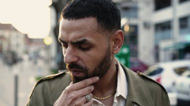 Düşünceli bir Arap genç adam ciddi bir ifadeyle şehir caddesinde duruyor. Şehir kaldırımında bir Orta Doğulu erkek.