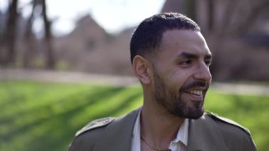 Güneşli bir günde parkta gülümseyen, kameraya doğru yürüyen mutlu bir Arap adam.