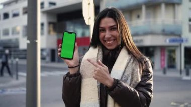 Çevrimiçi içeriği krom tuşlu yeşil ekran ile akıllı telefon aracılığıyla paylaşan mutlu genç bir kadın. Video sunumunda cep telefonu ekranına işaret eden kadın