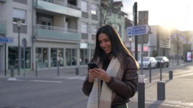 Gün batımında şehir caddesinde duran cep telefonu cihazına bakan mutlu bir genç kadın. 20 'li yaşlarda, elinde telefon tutan ve şehir ortamında gülümseyen yetişkin bir kız.
