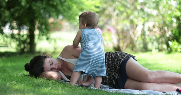 Tired mother trying to sleep outside laid on grass. Baby wanting mom attention, candid and authentic