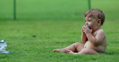 Baby infant seated on grass outdoors with ball inside mouth