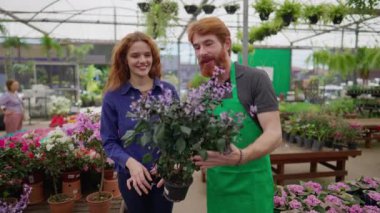 Yerel Çiçek Dükkanı 'nda bitki seçmeye yardım eden adam. Yeşil önlüklü kızıl saçlı bir erkek kadın müşteriye yardım ediyor.