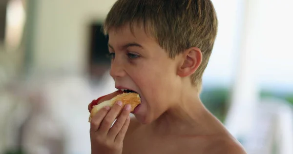 若い男の子の子供はホットドッグを一口食べる 子供がジャンクフードのスナックを食べる — ストック写真