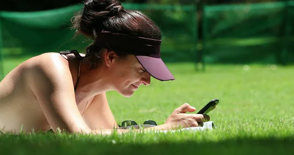 草の上に横になってスマートフォンを使っているカジュアルな女性 屋外の裏庭に携帯電話を入力する候補の女の子 — ストック写真