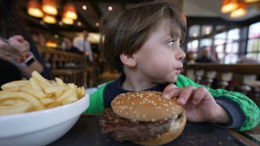 Restoranda oturmuş hamburger yiyen küçük bir çocuk. Düşünceli çocuk hamburger yiyor.