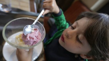 Küçük bir çocuk restoran masasında oturmuş kasede çilekli dondurma yiyor. Çocuk tatlı olarak kaşıkla yiyor.
