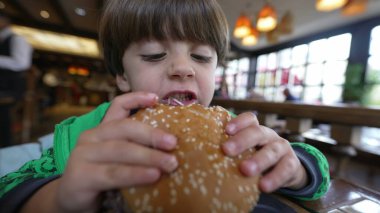 Hamburger yiyen genç bir çocuk. Çocuk hamburgerciden bir ısırık alıyor. Çocuk lokantada lezzetli yemekler yiyor.