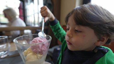 Dondurma yiyen küçük bir çocuk. Çocuk lezzetli çilek ve vanilyalı dondurma yiyor, kaşıkla restoranda oturuyor.