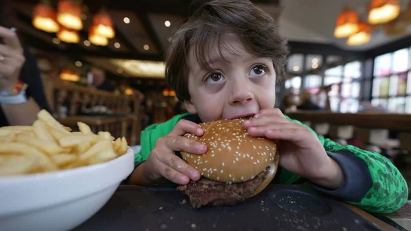 Restoranda oturmuş hamburger yiyen küçük bir çocuk. Düşünceli çocuk hamburger yiyor.