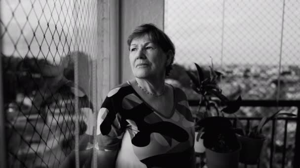 悲しい落ち込んでいるシニア女性だけで自宅の窓から市内の景色を深く調べるに立って感じている 高齢退職女性でモノクローム黒と白 — ストック動画