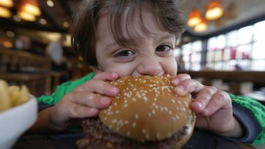Restoranda oturup hamburger yiyen aç küçük bir çocuk. Beyaz bir erkek çocuk hamburgerden bir ısırık alıyor. Çocuk lezzetli yemekler yiyor.