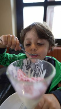 Neşeli çocuk cam kasede dondurma yiyor. Yakından bakınca çocuk tatlı yiyor. Yemekten sonra restoranda oturuyor.