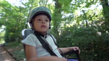 Bisikletli çocuk arka koltukta, kasklı, doğa yolunda gidiyor. Küçük çocuk ormanda annesiyle bisiklet sürüyor.