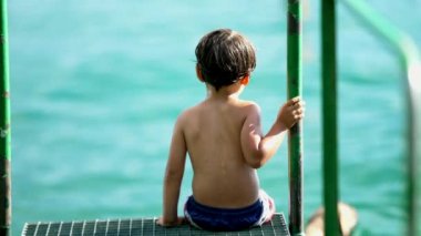 Metal çubuğa tutunarak suyun kenarında oturan düşünceli bir çocuk. Doğanın dışında derin düşüncelere dalmış dalgın küçük çocuk.