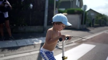 Güneşli bir günde şehrin yaya geçidinde üç tekerlekli scooter 'ı olan çocuk karşıya geçiyor. Şirin, aktif, üstsüz küçük çocuk caddeyi geçiyor.