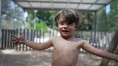 Dışarıda oynayan sevimli üstsüz çocuk kameraya gülümsüyor oyun parkında durup havaya çamur atıyor.