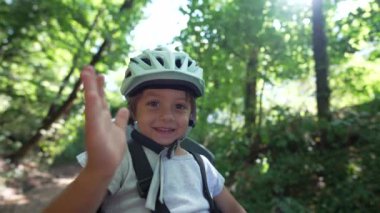 Bisiklet koltuğunda oturan ve kask takan mutlu bir çocuk el sallıyor. Bisiklet koltuğunda el sallayan sevimli çocuk.