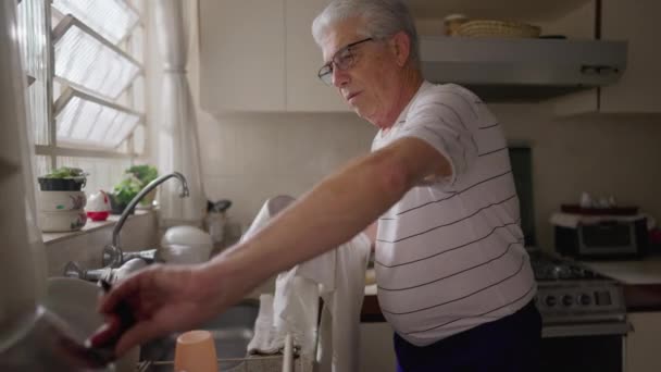 キッチンに立つシニア男性は自宅で窓ごと鍋やフライパンを整理シンクします 老人の本物の家庭シーン — ストック動画
