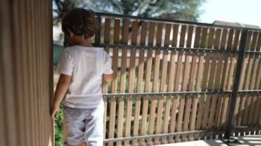 Balkon ahşap parmaklıklara tutunup apartmanın ikinci katından dışarı bakan mutlu küçük bir çocuk. Güneşli bir günde çocuk terasta dikilip dışarıya bakıyor.