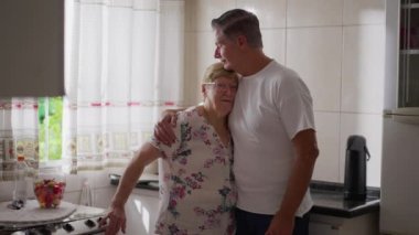 Yetişkin oğul, evde mutfakta dururken yaşlı anneyi kucaklıyor. Ev yaşamında özgün sevgi dolu bir aile anı.