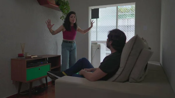 Couple in crisis discussing relationship. Emotionally upset girlfriend arguing with boyfriend while he is sitting on couch and she stands in disbelief
