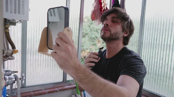 stock image One young man shaving beard with trimmer on the go while standing in apartment balcony and holding a mirror