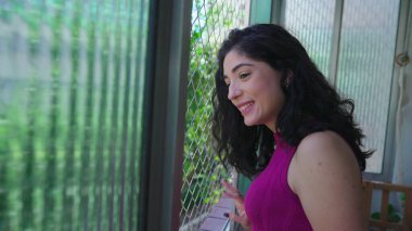 Mutlu, genç bir kadın daire penceresinden dışarı bakıp gülümsüyor. 30 'lu yaşlarda balkondan dışarı bakan neşeli bir kadın.