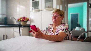 Ev mutfağında akıllı telefon kullanan yaşlı bir bayan, modern teknolojiyle meşgul olan ev içi yaşlılık yaşam tarzı.