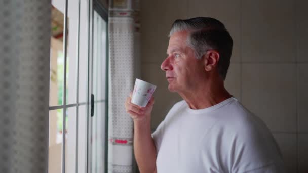 中年男人每天早晨都站在厨房窗边 一边喝着咖啡 一边喝着热饮着白发男人的日常生活方式 — 图库视频影像